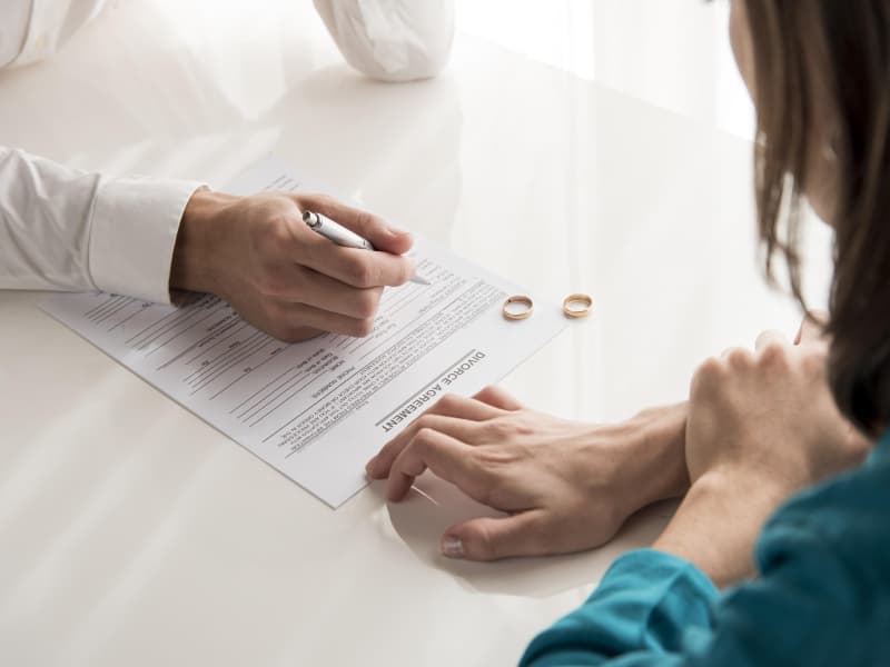Casal sentado à mesa assinando um documento, com duas alianças ao lado