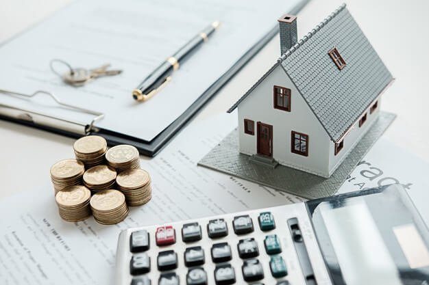 Maquete de casa sobre documentos, ao lado de moedas empilhadas e calculadora