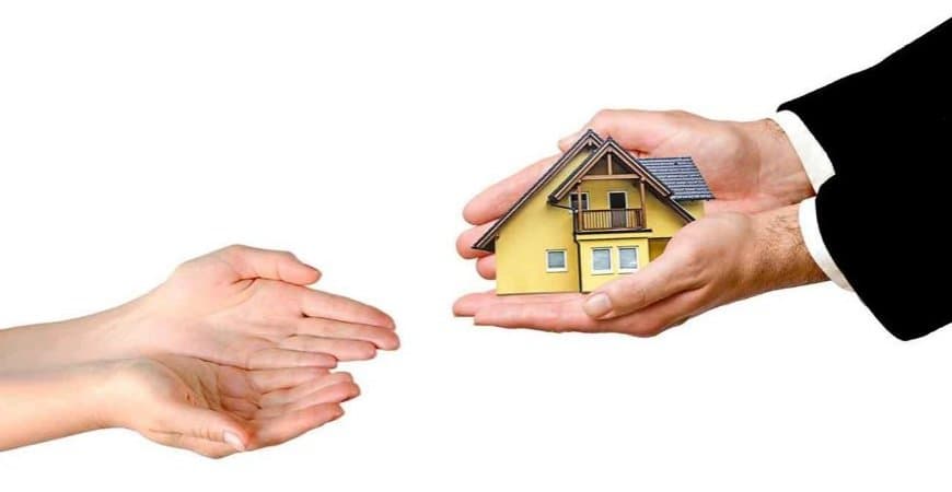 Mão de terno entregando a miniatura de uma casa para duas mãos abertas