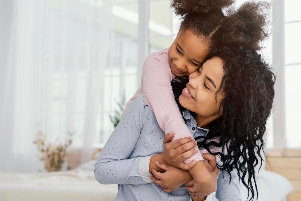 Mãe abraçada à filha pequena, que sorri apoiada em seu ombro