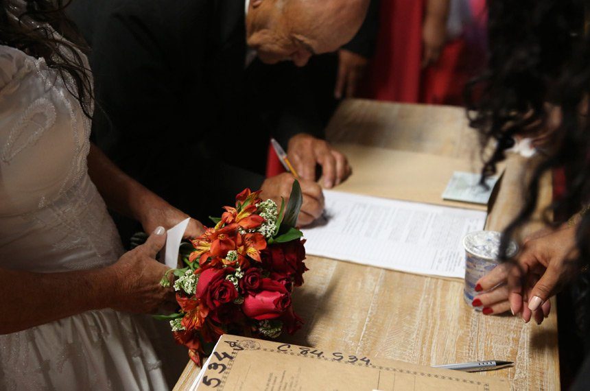Noivo assinando o documento de casamento no cartório, ao lado da noiva com buquê vermelho