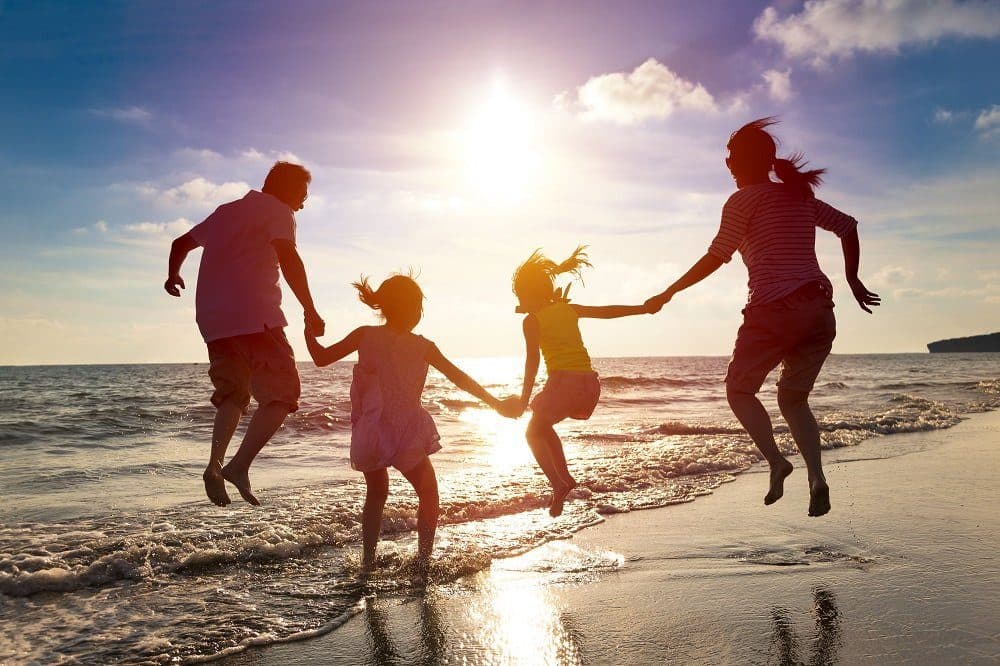 Família de quatro pessoas de mãos dadas, saltando na beira da praia ao pôr do sol